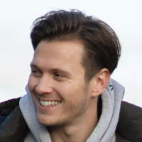 Smiling man with short dark hair wearing a gray hoodie and black jacket, outdoors.