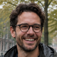 Close-up of smiling man with dark hair and glasses outdoors, trees in background.